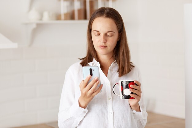 Mujer de cabello oscuro de pie con teléfono inteligente, leyendo noticias en las redes sociales, disfrutando de un café o té caliente en la cocina por la mañana, vestida con camisa blanca de estilo casual.