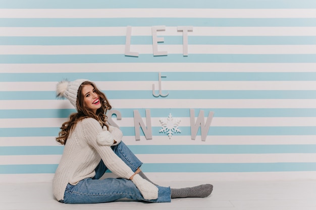 Mujer con cabello oscuro y rizado de buen humor posa sentada con elegante ropa de invierno contra la pared con letras Let it snow