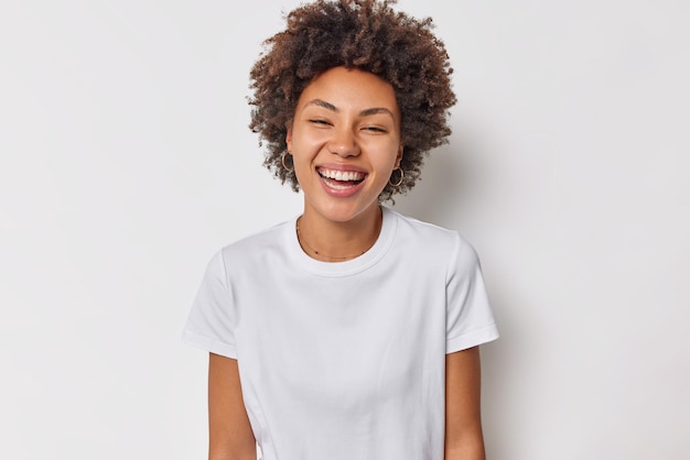 Mujer con cabello oscuro y rizado sonríe positivamente tiene un estado de ánimo optimista La expresión alegre viste una camiseta básica casual aislada en blanco. Sinceras emociones humanas.