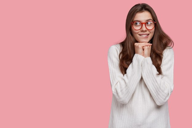 Mujer de cabello oscuro soñadora muerde los labios y mira a un lado, piensa en cosas agradables, mantiene las manos juntas