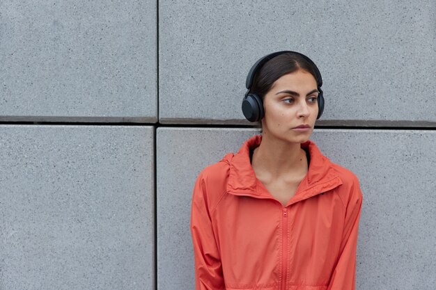 Mujer con cabello oscuro vestida con anorak casual mira hacia otro lado pensativamente escucha música a través de auriculares inalámbricos posa contra la pared gris espacio en blanco al aire libre para su información