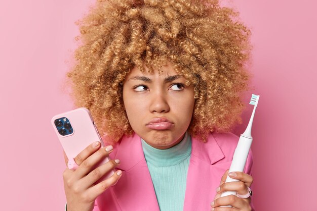 La mujer de cabello rizado descontenta tiene una expresión sombría que sostiene un teléfono móvil moderno y un cepillo de dientes eléctrico se somete a procedimientos de higiene vestidos con poses formales de chaqueta contra el fondo rosa del estudio