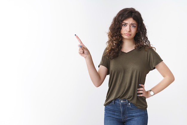 Mujer con cabello rizado frunciendo el ceño apuntando hacia la esquina superior izquierda cuestionada con desdén de pie vacilante expresar incredulidad sentimiento desagradable