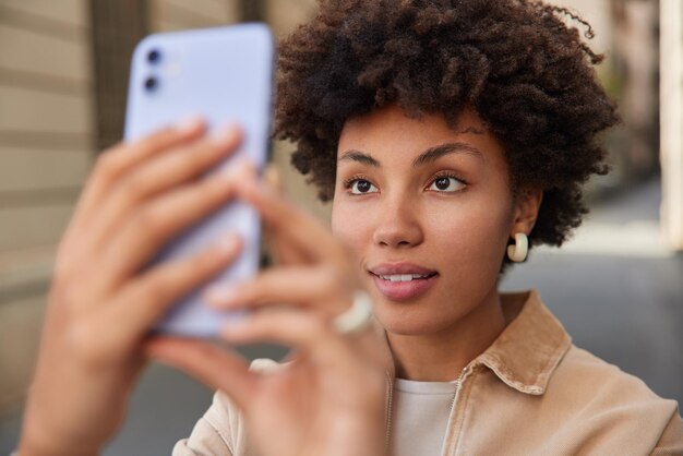 mujer con cabello rizado hace fotos en el dispositivo graba videos a través de teléfonos móviles digitales paseos en la calle viste ropa de moda enfocada en la pantalla disfruta de un día soleado