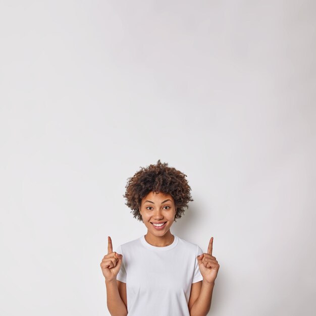 mujer con cabello rizado indica arriba con los dedos índices muestra la dirección o el lugar de su anuncio viste camiseta casual aislado en blanco. Concepto de publicidad