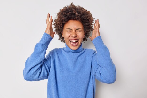 mujer con cabello rizado levanta las manos en la cabeza exclama de felicidad vestida con un jersey azul casual se siente feliz aislado en blanco. Concepto de emociones humanas