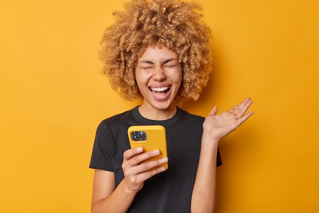 Una mujer de cabello rizado llena de alegría usa el teléfono móvil y se ríe de algo divertido que mantiene la palma levantada vestida con una camiseta negra aislada sobre un fondo amarillo chatea en línea Comunicación de tecnología de personas