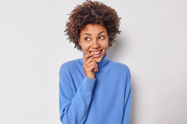 mujer con cabello rizado mantiene el dedo cerca de la boca sonríe con los dientes mira con admiración a un lado se siente tímida en presencia de un extraño usa poses casuales de jersey azul sobre blanco