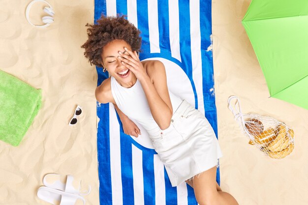 mujer con cabello rizado mantiene la mano en la cara sonríe alegremente vestida con camiseta blanca y falda posa sobre una toalla rayada disfruta del horario de verano pasa todo el día en la playa