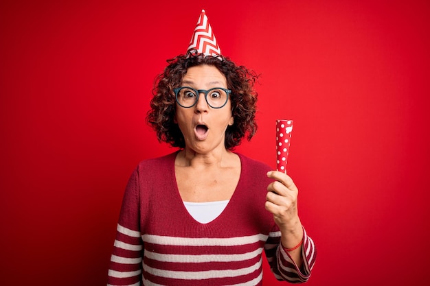 Mujer de cabello rizado de mediana edad con sombrero divertido de cumpleaños sosteniendo trompeta de fiesta en celebración asustada en shock con una cara sorpresa asustada y emocionada con expresión de miedo