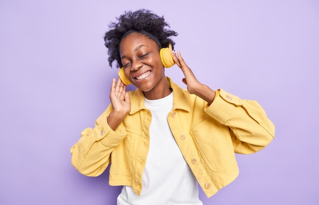 mujer con cabello rizado natural mantiene las manos en los auriculares relajados con la música siente el disfrute de las buenas canciones de audio usa una chaqueta amarilla aislada en la pared púrpura