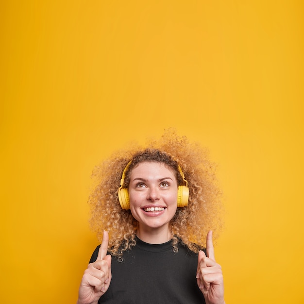 Mujer con cabello rizado natural sonríe positivamente muestra dientes blancos puntos arriba con los dedos índices tiene una expresión alegre escucha música a través de auriculares inalámbricos poses