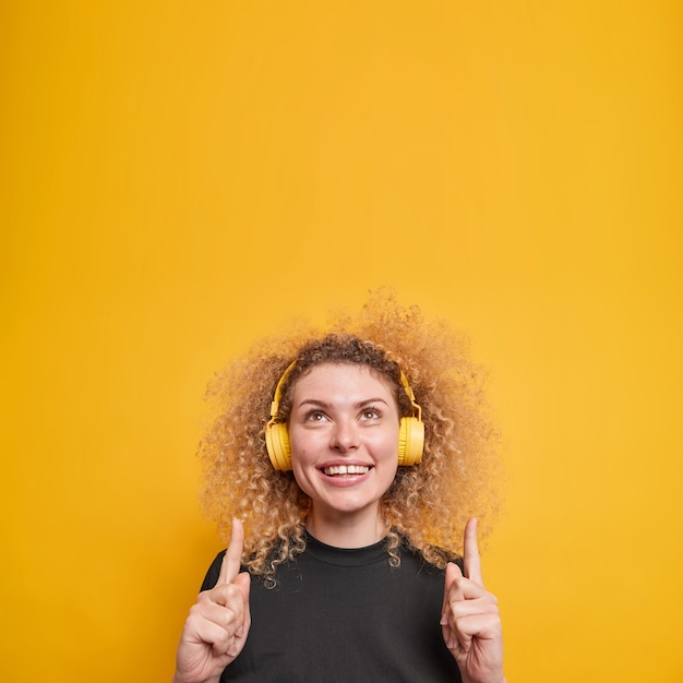 mujer con cabello rizado natural sonríe positivamente muestra dientes blancos puntos arriba con los dedos índices tiene una expresión alegre escucha música a través de auriculares inalámbricos poses