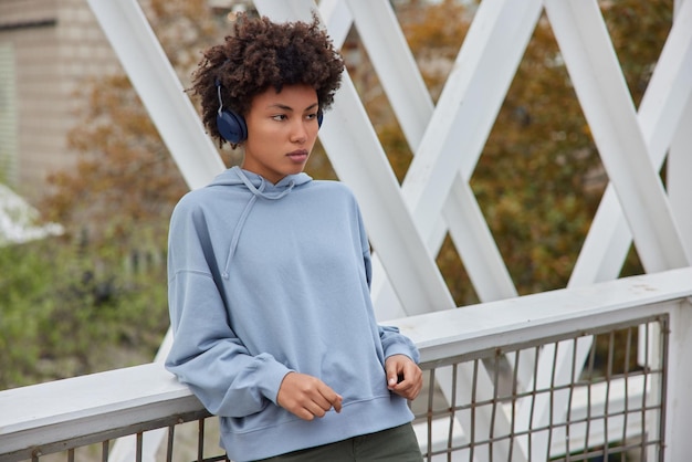 Mujer con cabello rizado posa en el puente viste una sudadera azul y pantalones escucha música motivadora a través de auriculares inalámbricos enfocados en la distancia. concepto de ocio y pasatiempo de personas.
