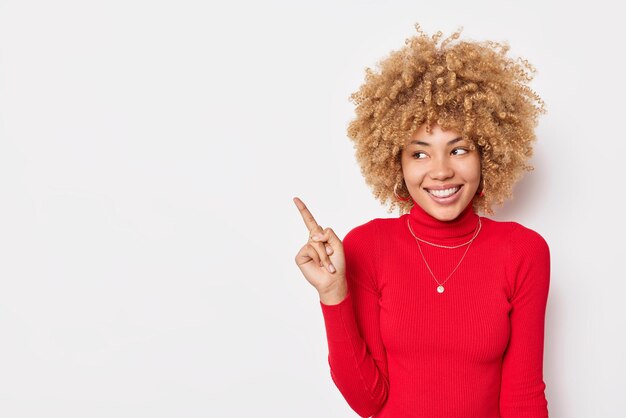 Una mujer de cabello rizado positivo señala con el dedo en el lado izquierdo y muestra un espacio en blanco para su contenido publicitario viste un cuello de tortuga rojo casual aislado sobre fondo blanco Coloque el título de su empresa aquí