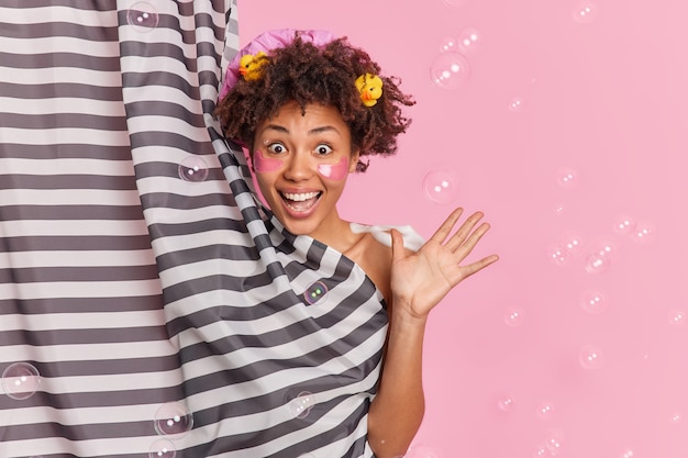 La mujer de cabello rizado refrescada positiva levanta la palma de la mano se siente muy feliz y emocionada se ducha se aplica parches de colágeno disfruta del cuidado de la piel y los procedimientos de higiene se divierten en el baño