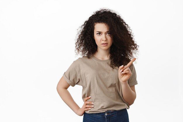 Mujer con cabello rizado sacudiendo el dedo, frunciendo el ceño y regañando a alguien que se porta mal, desaprueba algo, enseñando una lección, de pie sobre blanco