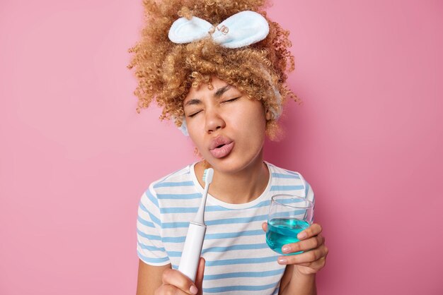 Una mujer de cabello rizado sostiene un cepillo eléctrico como si el micrófono cantara una canción sostiene un vaso de enjuague bucal usa una banda para la cabeza y una camiseta casual a rayas aislada sobre fondo rosa Concepto de higiene oral personal