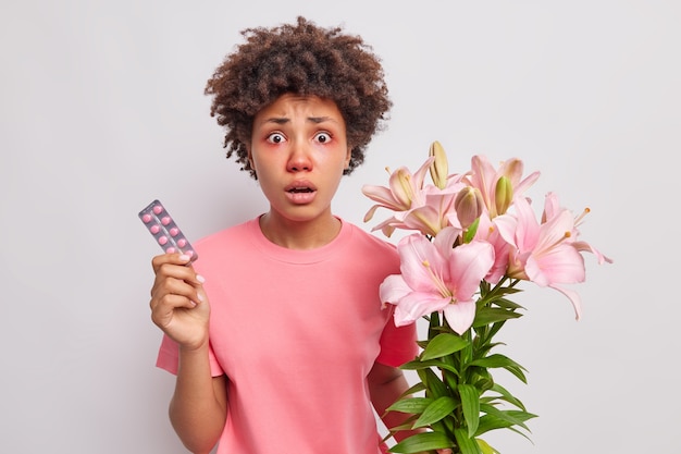 mujer con cabello rizado sostiene un ramo de flores de lirio tiene una reacción alérgica al polen sostiene medicamentos para curar los síntomas de la enfermedad usa una camiseta rosa aislada en blanco