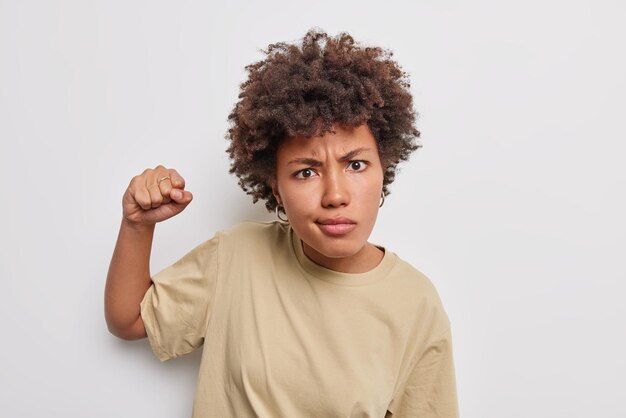 mujer con cabello rizado te advierte que la venganza levanta el puño cerrado mira con enojo viste una camiseta beige casual aislada en blanco. Si tan solo escucho esto una vez más