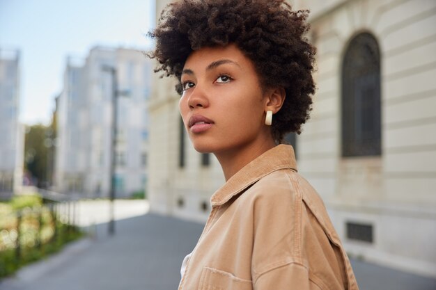 Mujer con cabello rizado y tupido mira a la distancia tiene un aspecto encantador vestido con una chaqueta beige pasea al aire libre espacio de copia en blanco