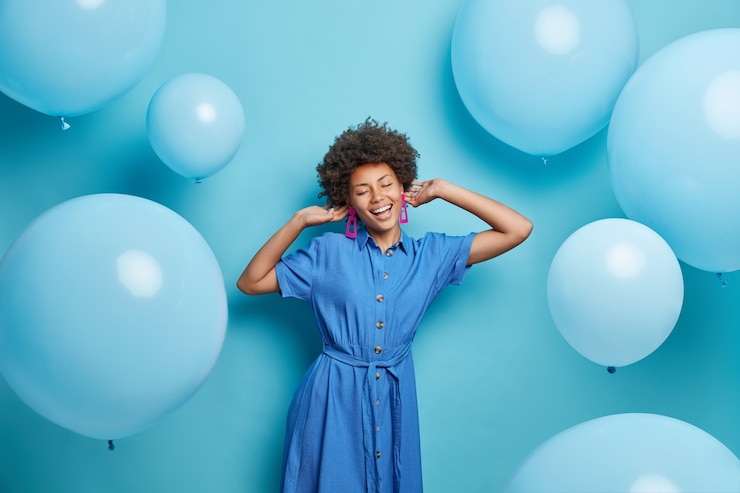 Foto gratuito mujer con cabello rizado vestida con un traje de moda disfruta de la música y poses de fiesta alrededor de globos inflados tiene moos festivos aislados en azul