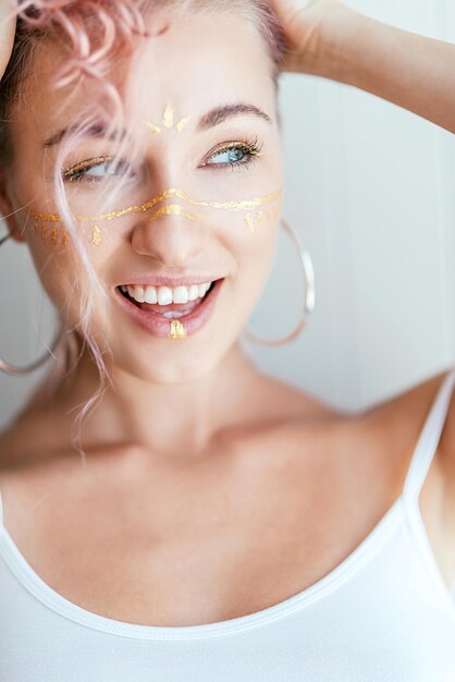 Mujer con cabello rosado y maquillaje artístico en forma de trazos de pintura sonriendo a un lado, posando en blanco claro