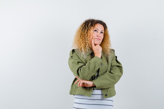 Mujer con cabello rubio rizado apoyando la barbilla en la mano en chaqueta verde y mirando pensativo, vista frontal.