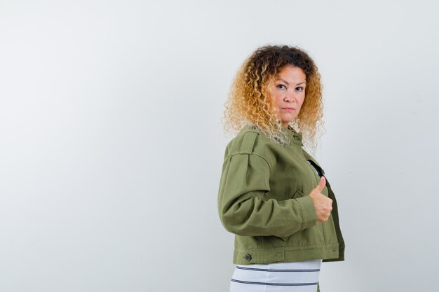 Mujer con cabello rubio rizado mostrando el pulgar hacia arriba en chaqueta verde y luciendo confiada. vista frontal.