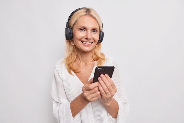 Mujer con cabello rubio sostiene teléfono móvil escucha música favorita de la lista de reproducción usa auriculares en las orejas usa blusa de moda aislada en blanco