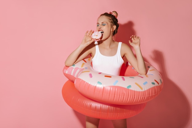 Mujer de cabello rubio en traje de baño blanco moderno comiendo donut y posando con dos grandes anillos de natación sobre fondo rosa aislado