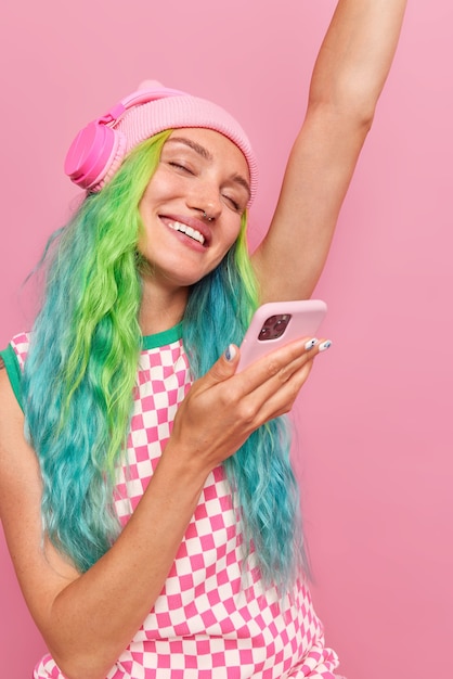 Mujer con cabello teñido de colores baila con el brazo levantado escucha música favorita a través de auriculares sostiene teléfono móvil aislado en rosa