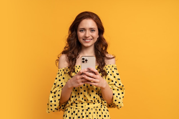 Mujer de cabeza roja sonriente dichosa que sostiene el teléfono de Mobyle en vestido amarillo del verano que presenta en amarillo