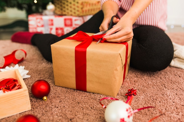 Mujer con caja de regalo y tijeras cerca de arcos decorativos, bolas y cinta en la alfombra