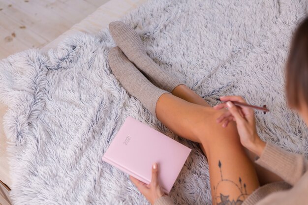 Mujer en calcetín de lana y suéter con letrero de cuaderno rosa 2021, gran tatuaje en la cadera. Mujer sentada en la cama en casa en el dormitorio.