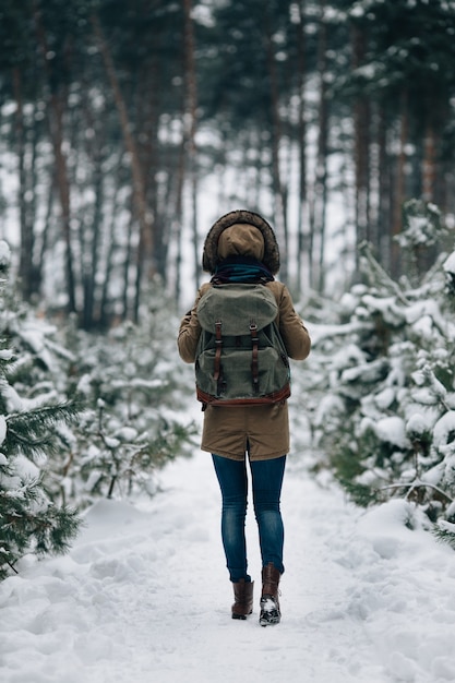 Mujer en cálida chaqueta de invierno con capucha de piel y mochila de viaje grande en el bosque de invierno cubierto de nieve