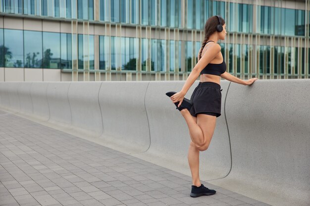 mujer se calienta antes de trotar levanta la pierna estira los músculos vestidos con ropa deportiva se prepara para el entrenamiento cardiovascular posa hacia adelante enfocada cerca del edificio de cristal de la ciudad moderna