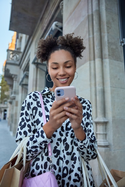 Mujer camina afuera con bolsas de papel, mensajes a través de un teléfono inteligente, feliz de recibir comentarios bajo su comentario, feliz de hacer compras exitosas durante el tiempo libre