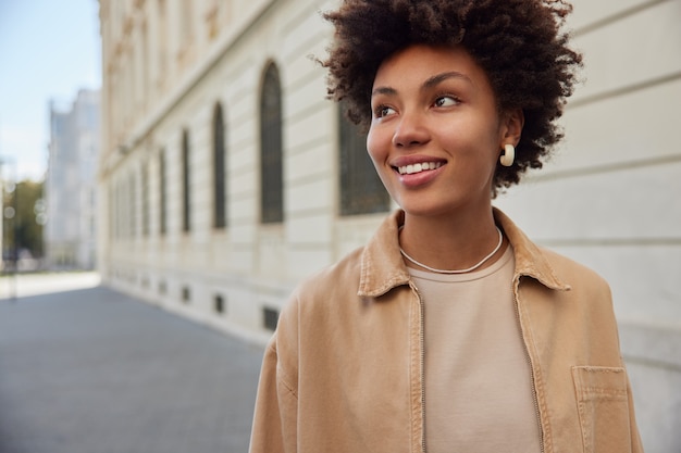 mujer camina al aire libre pasea en una pequeña ciudad hermosa alrededor de edificios antiguos sonríe felizmente viste ropa elegante disfruta del tiempo libre en vacaciones tiene expresión feliz