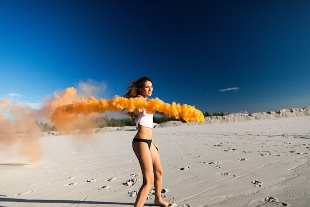 Mujer camina con humo de color naranja en la playa blanca bajo el cielo azul