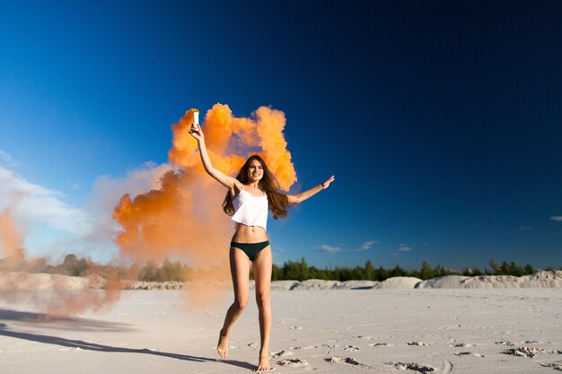 Mujer camina con humo de color naranja en la playa blanca bajo el cielo azul