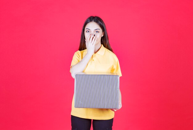 mujer con camisa amarilla sosteniendo una caja de regalo plateada y parece confundida y pensativa.