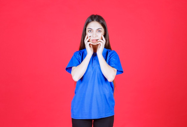 mujer con camisa azul de pie en la pared roja y se ve emocionada y aterrorizada.