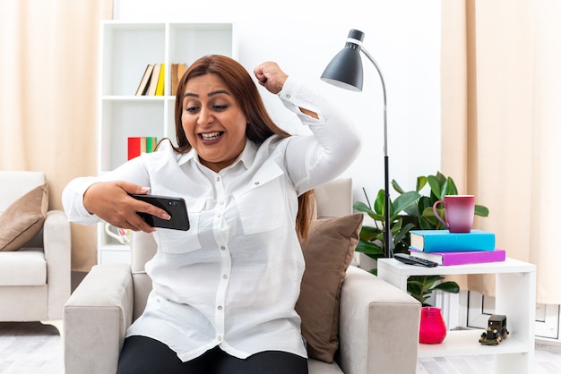 Mujer con camisa blanca y pantalón negro sosteniendo un teléfono inteligente feliz y emocionado levantando el puño regocijándose de su éxito sentada en la silla en la sala de estar luminosa