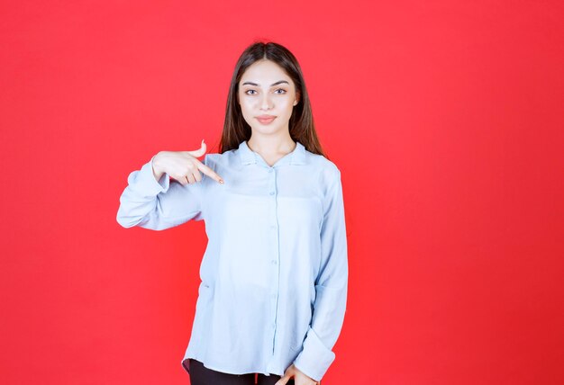 mujer con camisa blanca de pie sobre la pared roja y apuntando a sí misma.