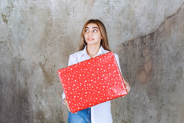 mujer con camisa blanca sosteniendo una caja de regalo roja con puntos blancos y se ve confundida y pensativa.