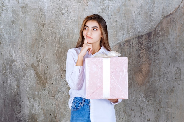 mujer con camisa blanca sosteniendo una caja de regalo rosa envuelta con una cinta blanca y parece confundida y vacilante.