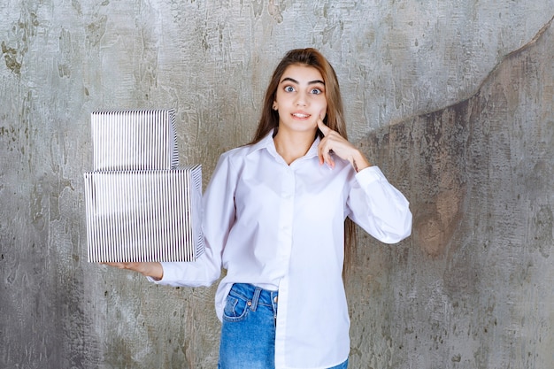 Mujer con camisa blanca sosteniendo cajas de regalo plateadas y parece confundida