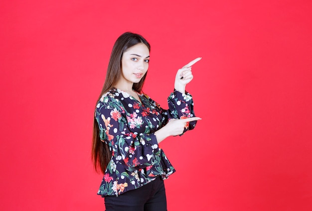 mujer con camisa floral de pie sobre la pared roja y apuntando a la derecha.