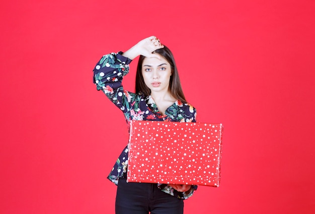 mujer con camisa floral sosteniendo una caja de regalo roja con puntos blancos y se ve confundida y pensativa.